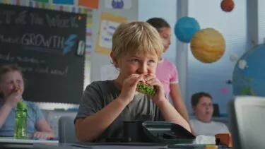 En ung dreng sidder ved et skrivebord i et klasseværelse og spiser en sandwich. Bag ham er der andre børn, og i baggrunden hænger farverige pædagogiske dekorationer og planetmodeller.