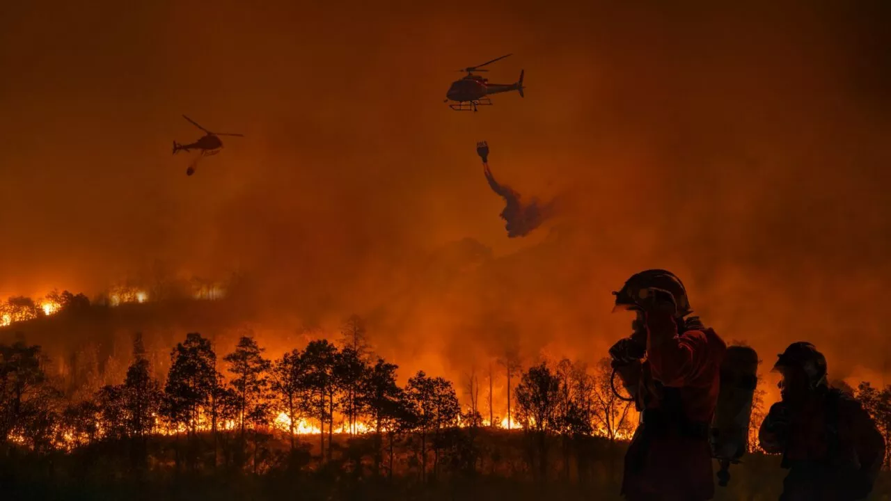 En stor skovbrand opsluger en skov om natten. To helikoptere kaster vand på flammerne, mens to brandmænd i beskyttelsesudstyr står i forgrunden, oplyst af den intense ild. Røg og orange skær fylder himlen.