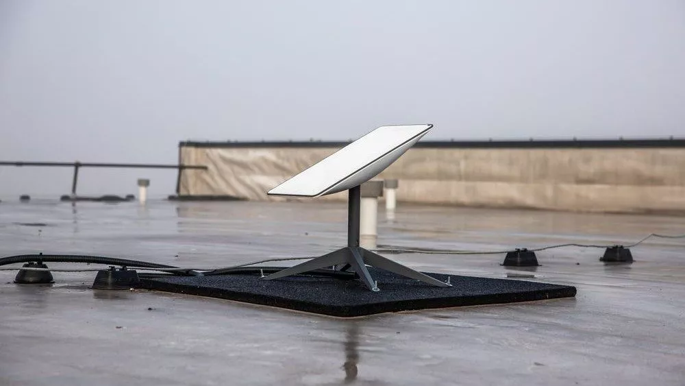 En Starlink-parabol sidder på en platform på taget med fastgjorte kabler, omgivet af en flad betonoverflade og en tåget baggrund.