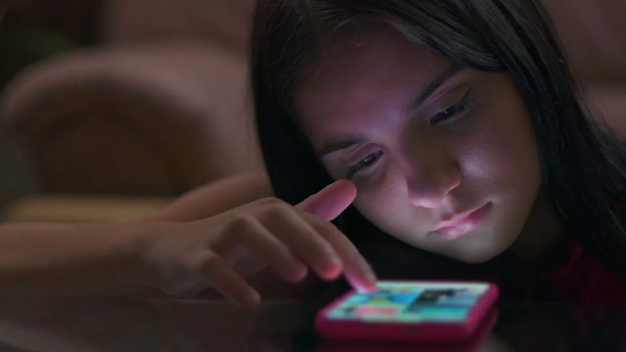 En ung pige med langt mørkt hår bruger intenst en smartphone med et pink etui, hendes ansigt oplyses af skærmen, mens hun rører ved displayet med sin finger.