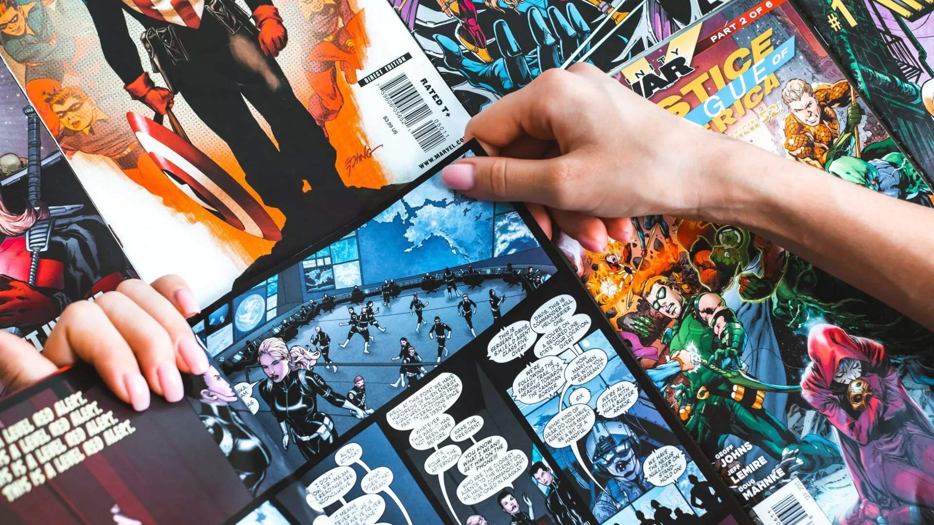 To hænder holder en tegneserie åben, omgivet af flere farverige Marvel-tegneserier, der ligger spredt på et bord. Den åbne tegneserie viser illustrerede figurer og talebobler.