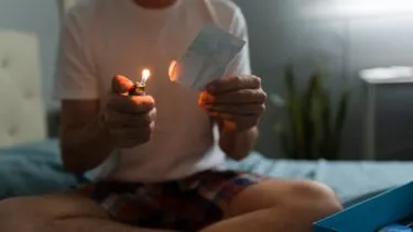En mand sidder med benene over kors på en seng og bruger en lighter til at brænde et fotografi, så den ene kant bliver delvist brændt. Han er iført en hvid t-shirt og ternede shorts og har en blå kasse ved siden af sig i et svagt oplyst rum.