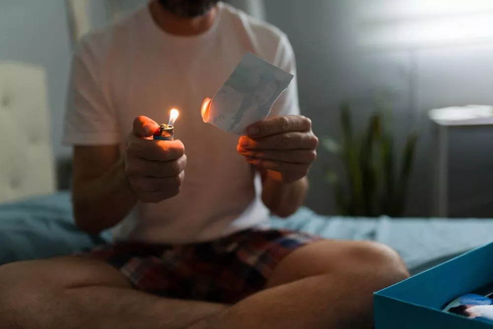 En mand sidder med benene over kors på en seng og bruger en lighter til at brænde et fotografi, så den ene kant bliver delvist brændt. Han er iført en hvid t-shirt og ternede shorts og har en blå kasse ved siden af sig i et svagt oplyst rum.