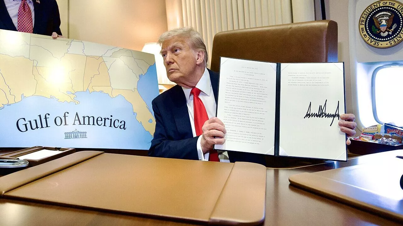En mand i jakkesæt sidder ved et skrivebord og holder et underskrevet dokument op. Bag ham holder en anden person et kort med påskriften Gulf of America. Det ser ud til, at de befinder sig om bord på et officielt regeringsfly.