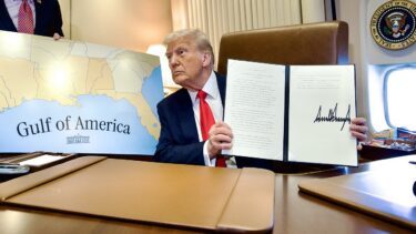 En mand i jakkesæt sidder ved et skrivebord og holder et underskrevet dokument op. Bag ham holder en anden person et kort med påskriften Gulf of America. Det ser ud til, at de befinder sig om bord på et officielt regeringsfly.