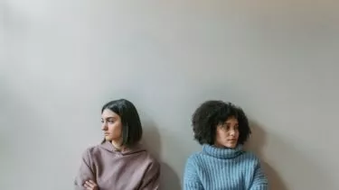 To kvinder sidder hver for sig op ad en almindelig væg, begge med armene over kors og kigger væk fra hinanden og virker fjerne eller oprørte.