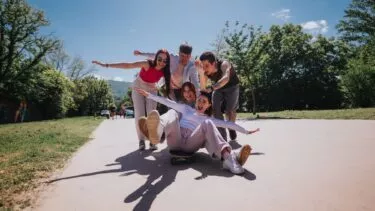 En gruppe på fem unge venner nyder en solrig dag udendørs; en sidder på et skateboard med udstrakte arme, mens de andre læner sig ind, smiler og griner sammen på en asfalteret sti omgivet af træer.