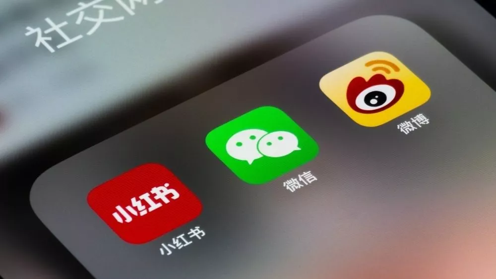 Nærbillede af en smartphone-skærm med tre kinesiske app-ikoner for sociale medier: Xiaohongshu (rødt ikon), WeChat (grønt ikon) og Weibo (gult ikon) på en mørk baggrund.