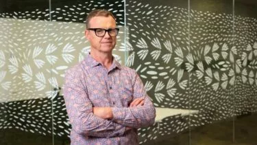 En mand med briller og mønstret skjorte står med korslagte arme foran en glasvæg dekoreret med et abstrakt, hvidt bladdesign.