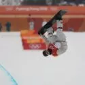 En snowboarder iført rød bib og hjelm udfører et upside-down aerial trick på en snedækket halfpipe ved vinter-OL, med Pyeongchang 2018-banneret synligt i baggrunden.