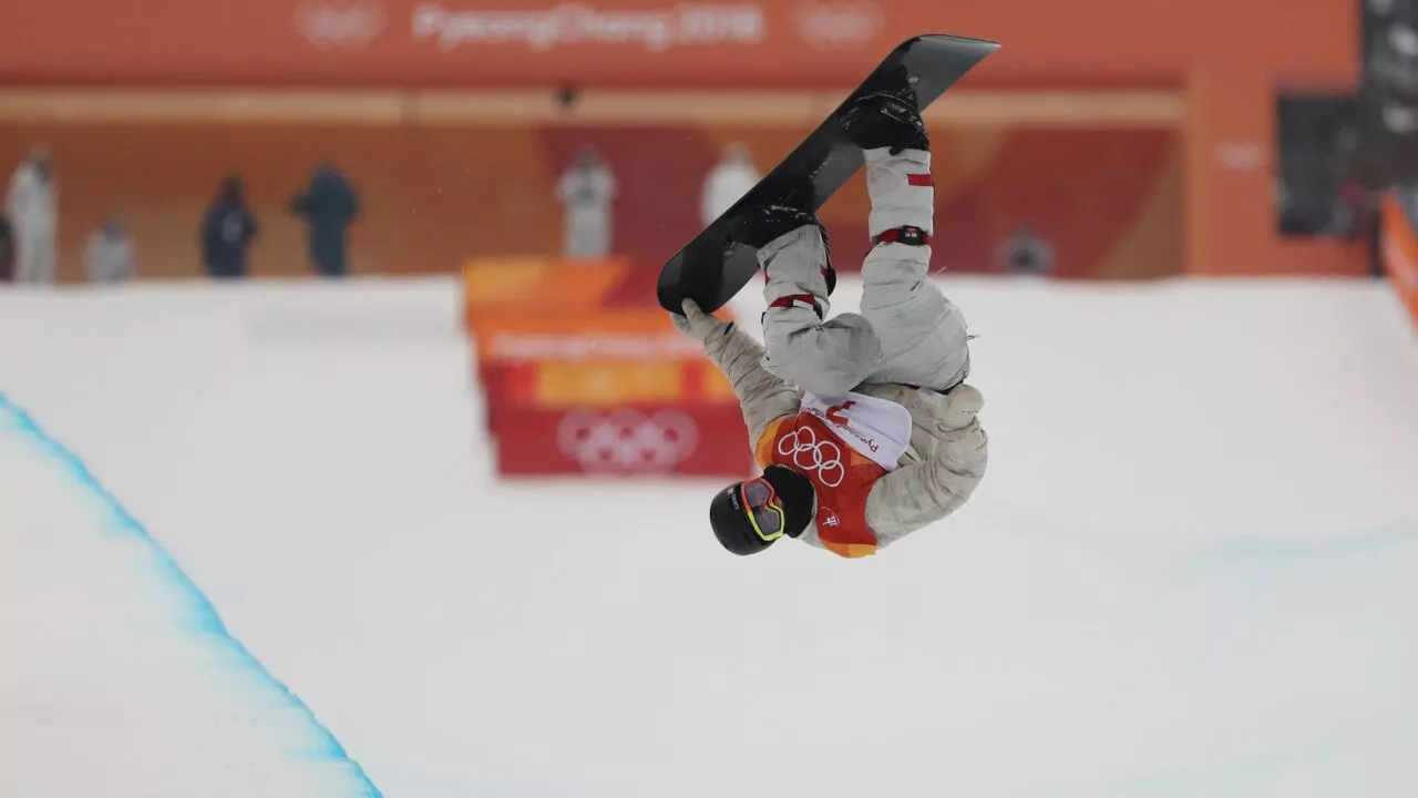 En snowboarder iført rød bib og hjelm udfører et upside-down aerial trick på en snedækket halfpipe ved vinter-OL, med Pyeongchang 2018-banneret synligt i baggrunden.