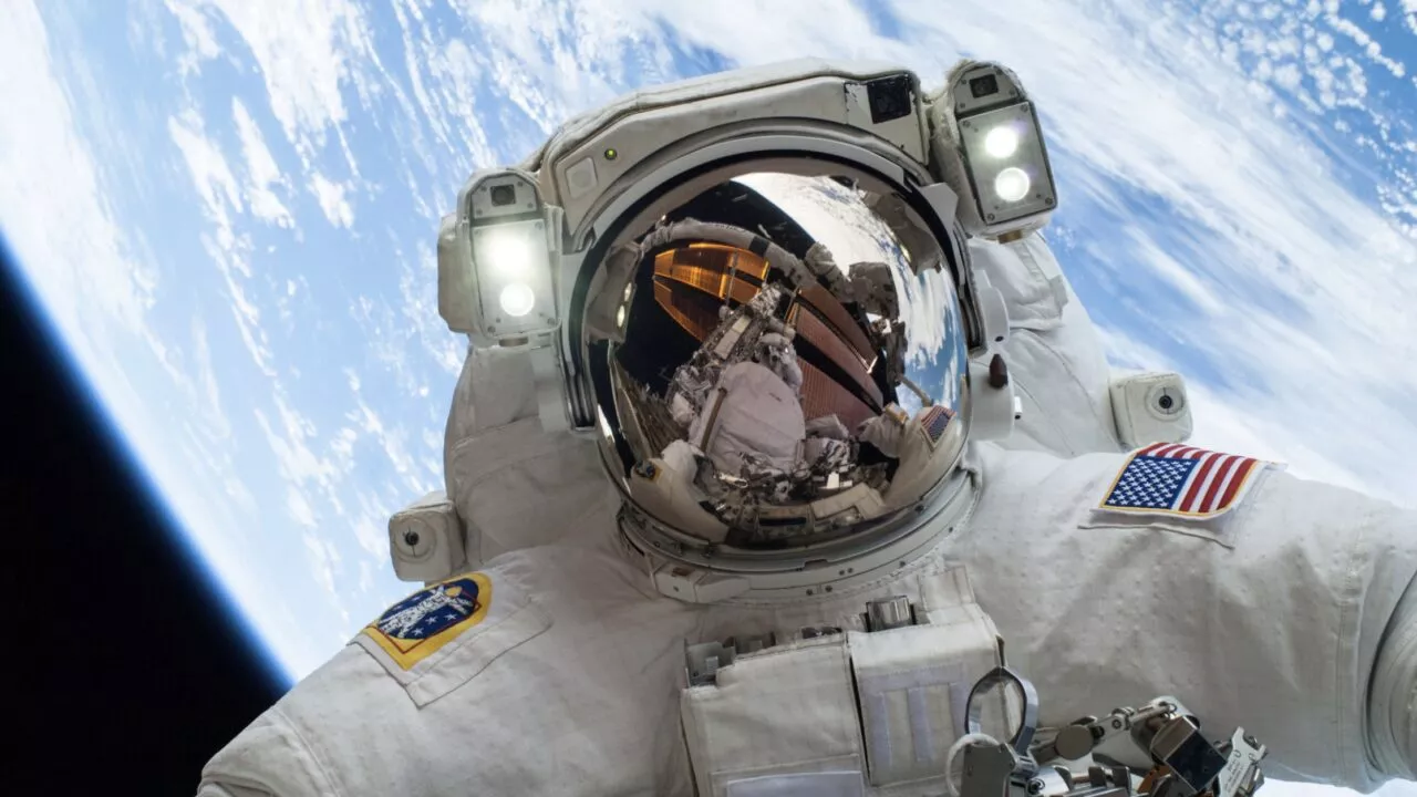 En astronaut i en hvid rumdragt svæver i rummet over Jorden, mens hjelmens visir reflekterer rumfartøjet og det omgivende udstyr. Planetens blå og hvide kurve er synlig i baggrunden.