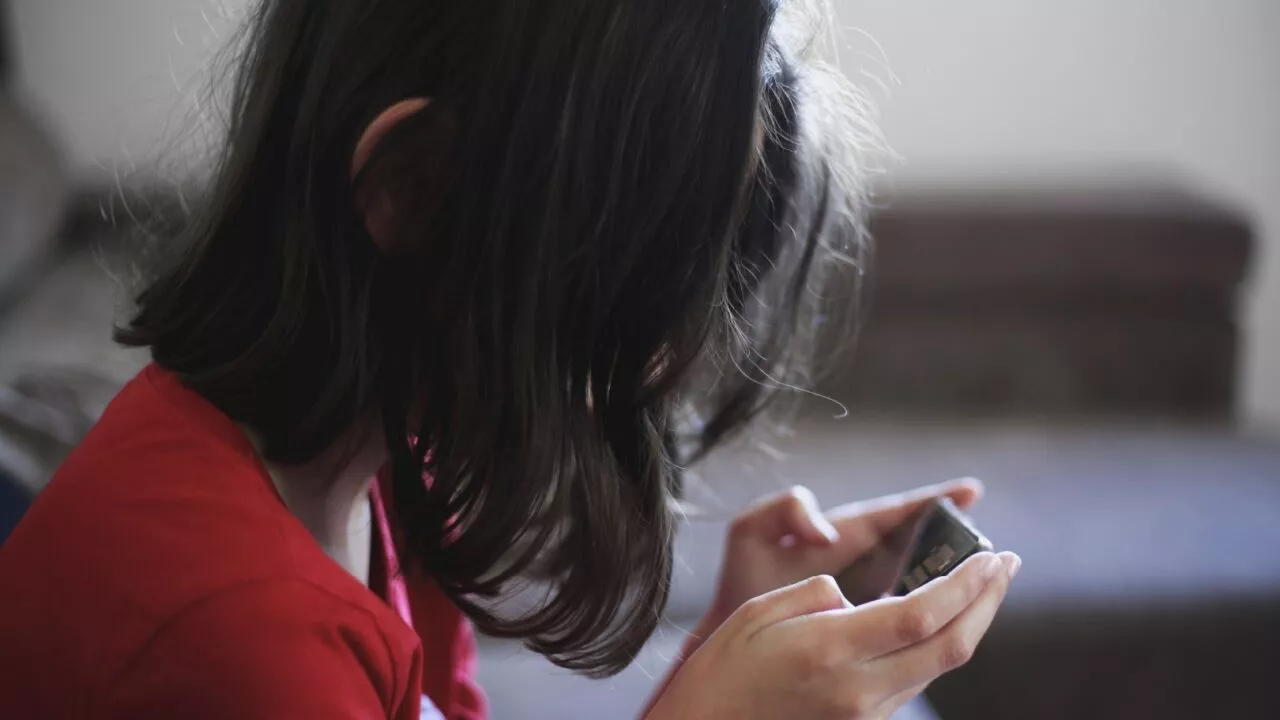 Et barn med skulderlangt mørkt hår iført en rød skjorte sidder indendørs og kigger ned, mens det holder og bruger en smartphone med begge hænder. Baggrunden er let sløret.