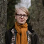 En ung person med lyst hår og briller står udendørs og smiler. De bærer et sennepsgult tørklæde, en ternet skjorte og en mørk jakke med træstammer og sløret grønt i baggrunden.