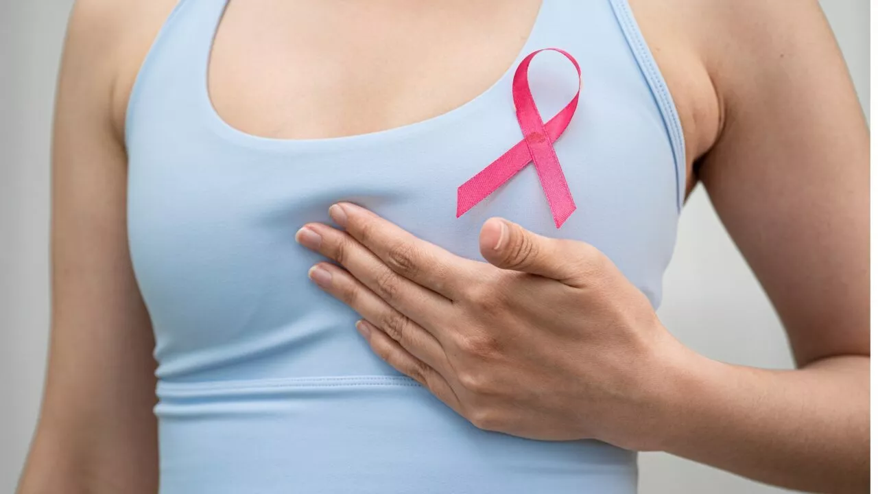 En person i en lyseblå tanktop rører ved sit bryst i nærheden af en pink sløjfe, der er sat fast på trøjen, og som symboliserer bevidsthed om brystkræft.