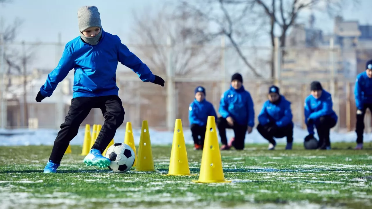 Et barn i blå jakke og grå hue dribler en fodbold gennem gule kegler på en græsplæne, mens andre børn i blå jakker ser til i baggrunden. Der er lidt sne på jorden.