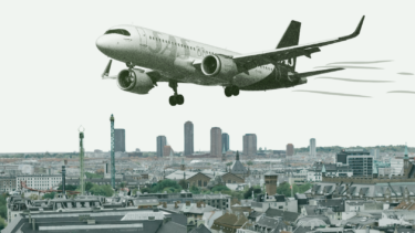 Et kommercielt fly flyver lavt over et bybillede med høje bygninger og hustage og ser ud til at forberede sig på at lande mod en lys himmel.