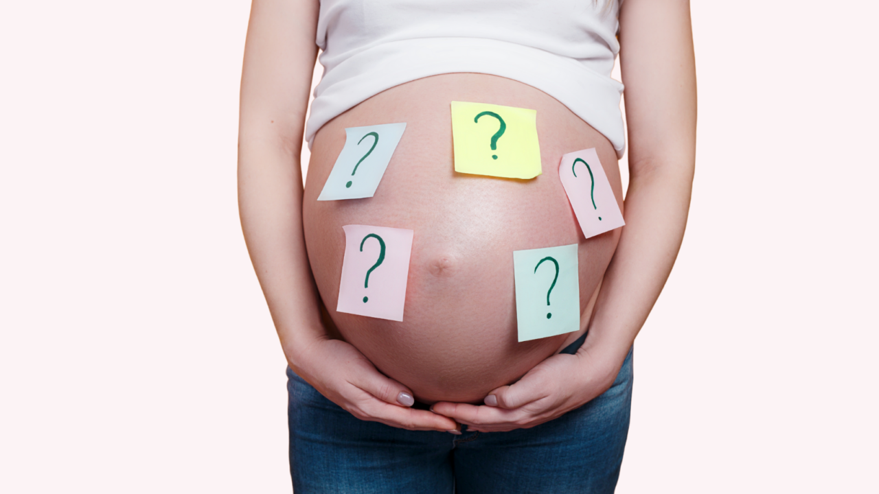 En gravid person iført jeans og hvid skjorte holder om sin mave, som er dækket af farverige klistermærker med spørgsmålstegn.
