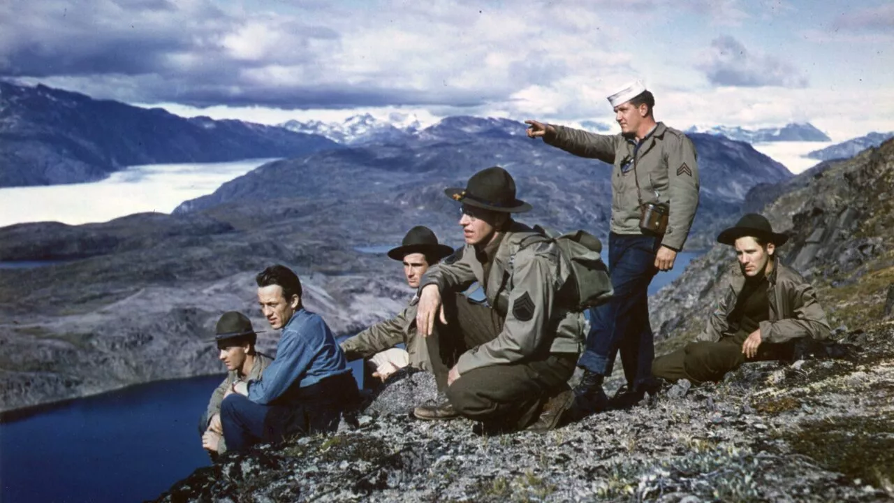 En gruppe soldater og en sømand i uniform sidder og knæler på en klippeskråning med udsigt over et bjergrigt landskab med en sø og en overskyet himmel i det fjerne. En mand står og peger ud mod horisonten.