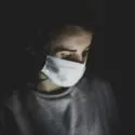 En person med hvid ansigtsmaske ser nedad i dæmpet belysning med bløde skygger, der fremhæver ansigtet og overkroppen mod en mørk baggrund.