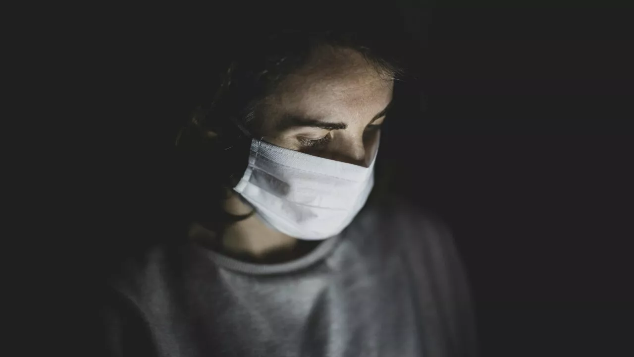 En person med hvid ansigtsmaske ser nedad i dæmpet belysning med bløde skygger, der fremhæver ansigtet og overkroppen mod en mørk baggrund.