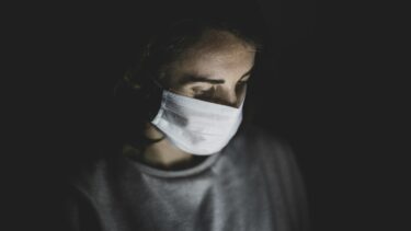 En person med hvid ansigtsmaske ser nedad i dæmpet belysning med bløde skygger, der fremhæver ansigtet og overkroppen mod en mørk baggrund.