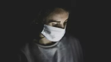 En person med hvid ansigtsmaske ser nedad i dæmpet belysning med bløde skygger, der fremhæver ansigtet og overkroppen mod en mørk baggrund.