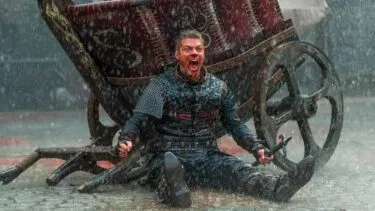 En hidsig mand i middelalderlig rustning sidder på mudret jord i kraftig regn, skrigende af raseri, med to våben i hånden, foran en stor trækvogn med et rødt og guldmønstret klæde.