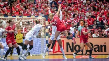 En håndboldkamp for kvinder er i gang, med spillere i røde og hvide uniformer, der hopper og rækker ud efter bolden nær målet, og en flok tilskuere i rødt, der ser på i baggrunden.