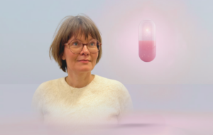 En kvinde med kort brunt hår og briller iført en cremefarvet sweater står foran en pastelfarvet baggrund med en stor, lysende pink kapsel svævende ved siden af sig.