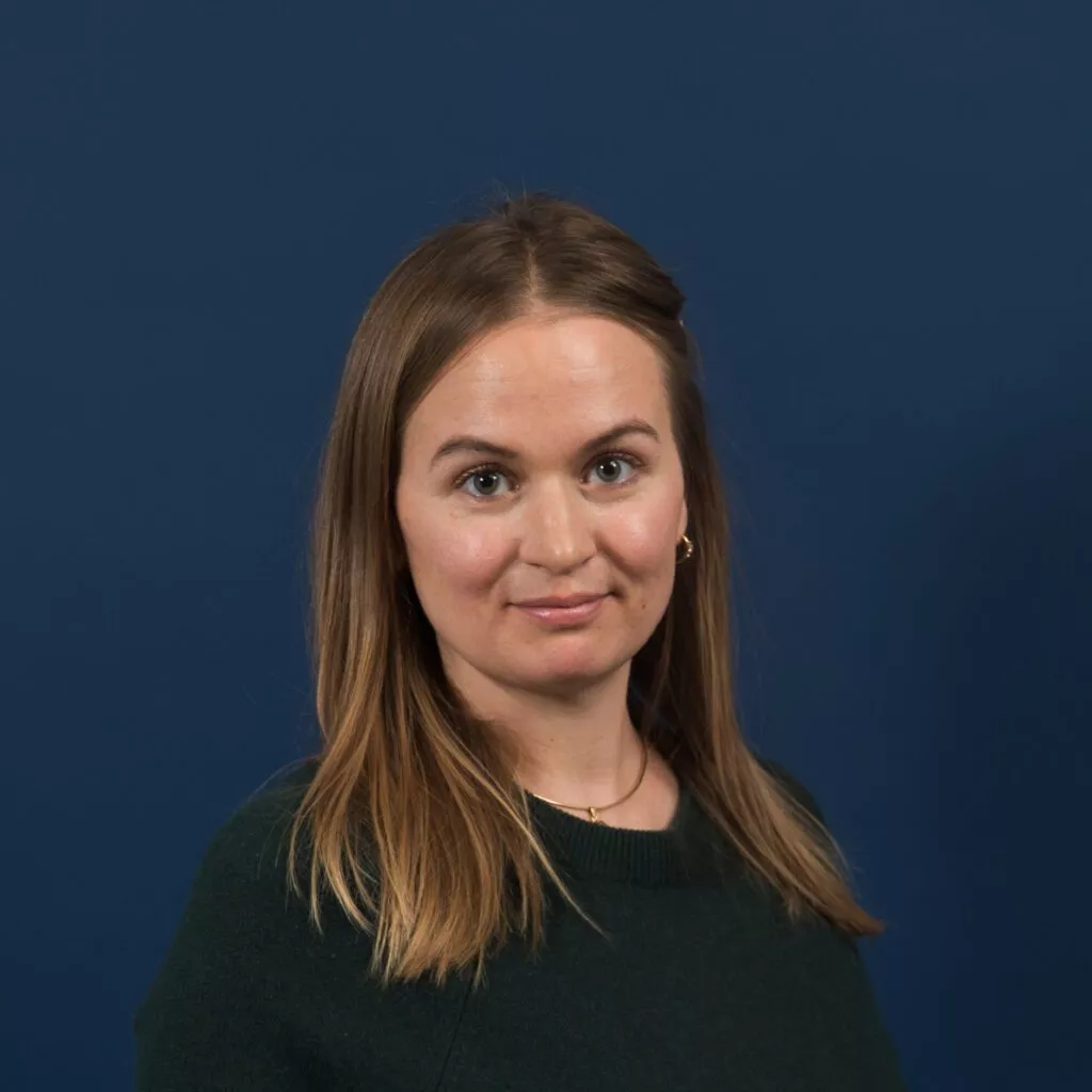En kvinde med glat, lysebrunt hår iført en mørkegrøn top står foran en almindelig mørkeblå baggrund og ser på kameraet med et neutralt udtryk.