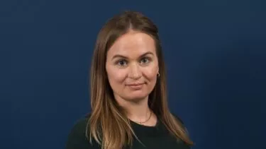 En kvinde med glat, lysebrunt hår iført en mørkegrøn top står foran en almindelig mørkeblå baggrund og ser på kameraet med et neutralt udtryk.