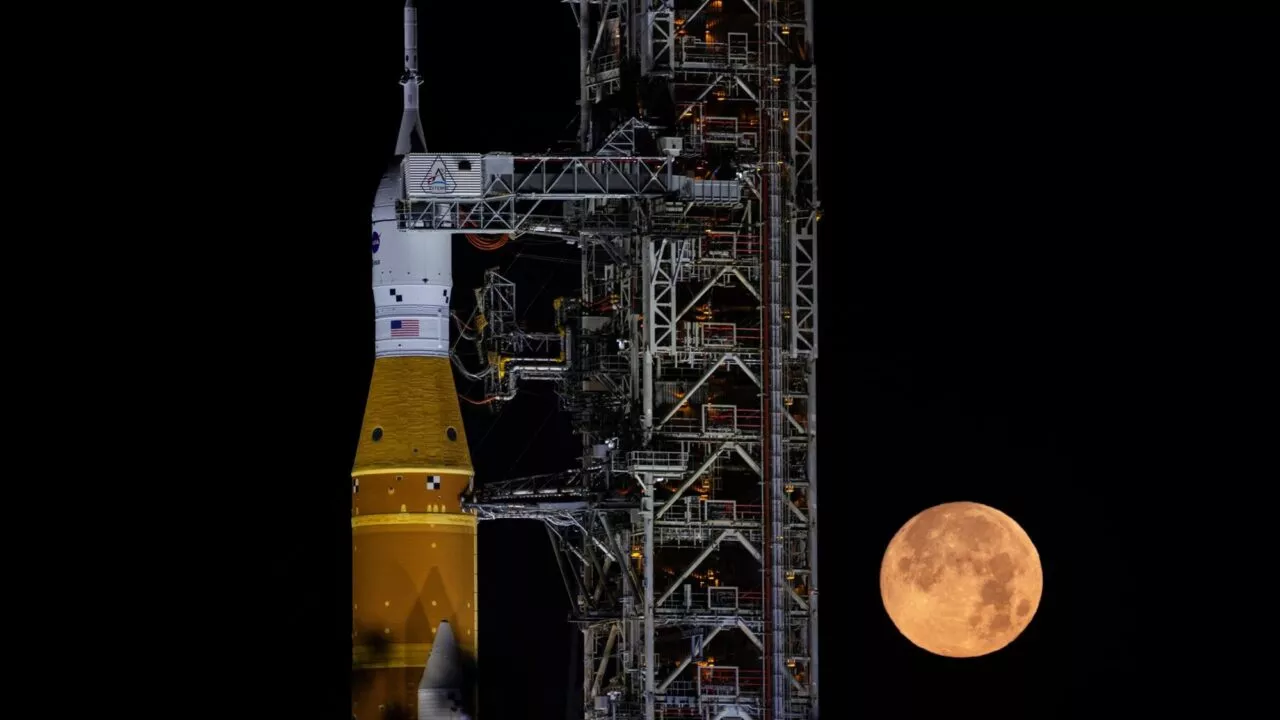 En NASA-raket står på en affyringsrampe om natten, oplyst mod en mørk himmel. En stor, lysende fuldmåne står lavt på himlen ved siden af affyringsanlægget.