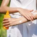 En person i hvidt tøj smører sin arm ind i solcreme udendørs, mens han holder en orange flaske. Baggrunden er grøn og sløret, hvilket antyder en solrig dag i naturen.
