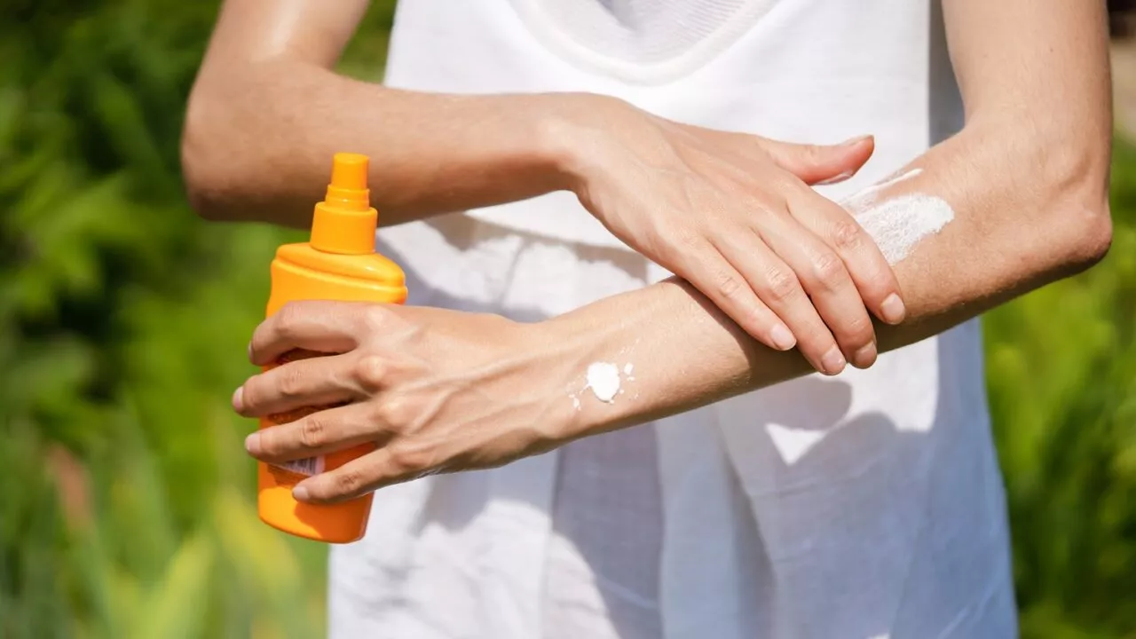 En person i hvidt tøj smører sin arm ind i solcreme udendørs, mens han holder en orange flaske. Baggrunden er grøn og sløret, hvilket antyder en solrig dag i naturen.