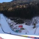 En snedækket skihopbakke med flere ramper, omgivet af grantræer og bjerge. Der er sponsorbannere langs pisten, og området er badet i vintersollys.