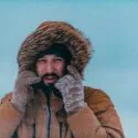 En person iført pelsbeklædt hætte og handsker står i snevejr med frost på tøj og hænder, rører ved sit ansigt og kigger ud i det fjerne mod en kold, blå himmel.