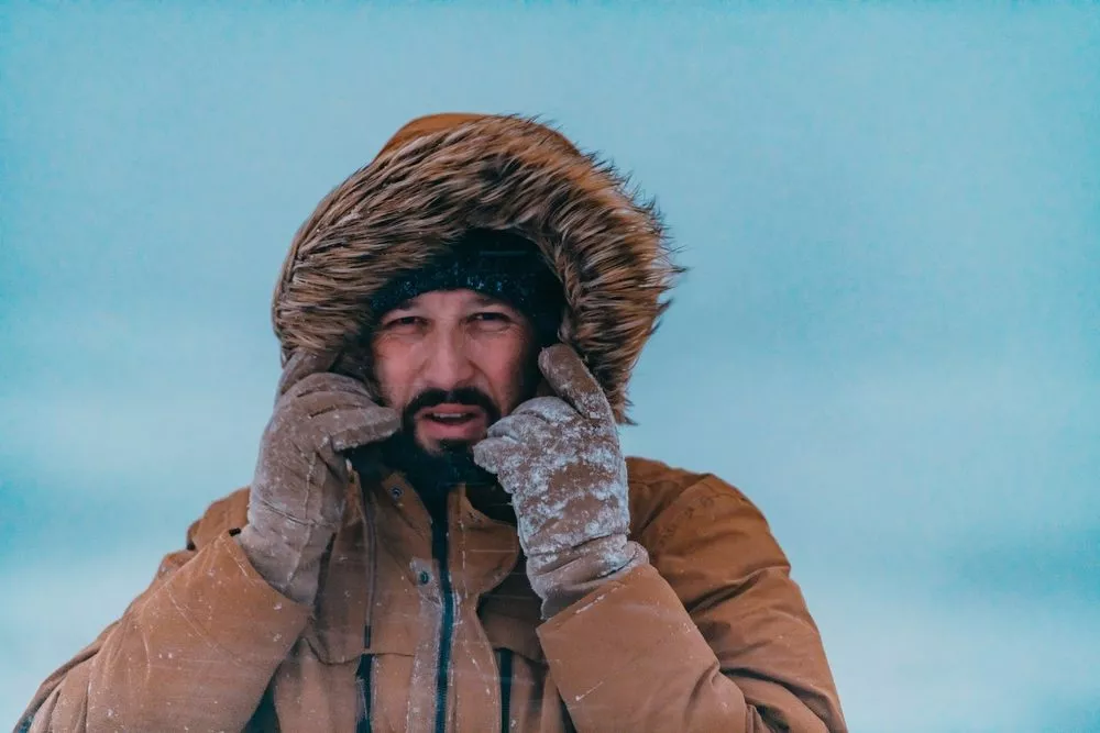 En person iført pelsbeklædt hætte og handsker står i snevejr med frost på tøj og hænder, rører ved sit ansigt og kigger ud i det fjerne mod en kold, blå himmel.