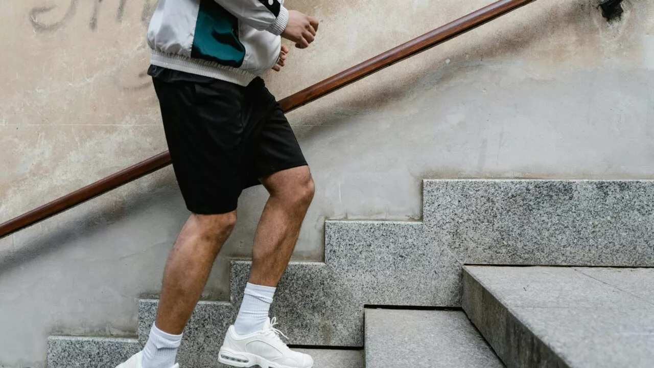 En person iført en lys jakke, sorte shorts, hvide sokker og hvide sneakers går op ad en stentrappe med den ene hånd på trægelænderet. Vedkommendes ansigt er ikke synligt.