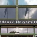 Skiltet Syddansk Universitet står på en moderne bygning med store glasvinduer, der reflekterer himlen og flagene.