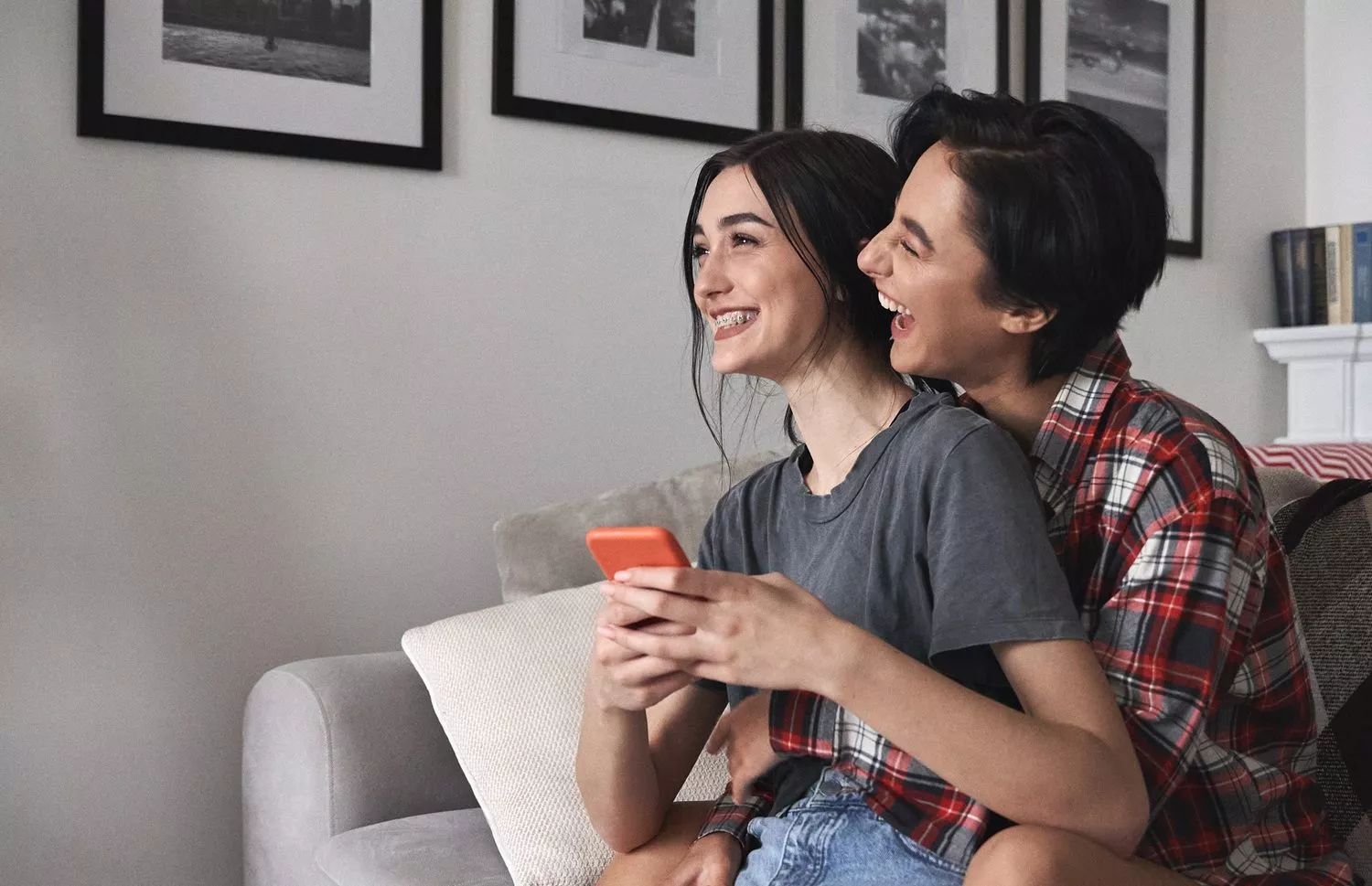 To mennesker sidder tæt sammen i en sofa og smiler og griner. Den ene holder en telefon, mens den anden krammer dem bagfra. Indrammede sort-hvide fotos hænger på væggen i baggrunden.