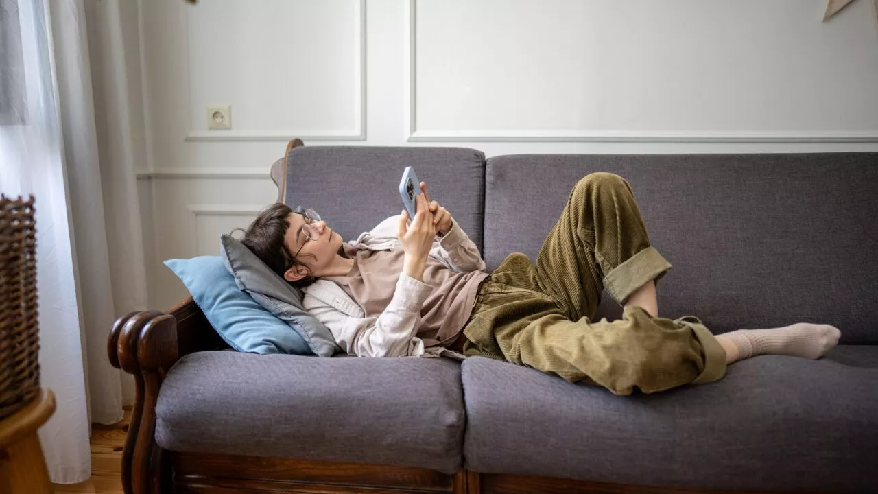 En person med briller og afslappet tøj ligger på en grå sofa, holder og kigger på en smartphone med hovedet hvilende på en pude i en hyggelig, veloplyst stue.