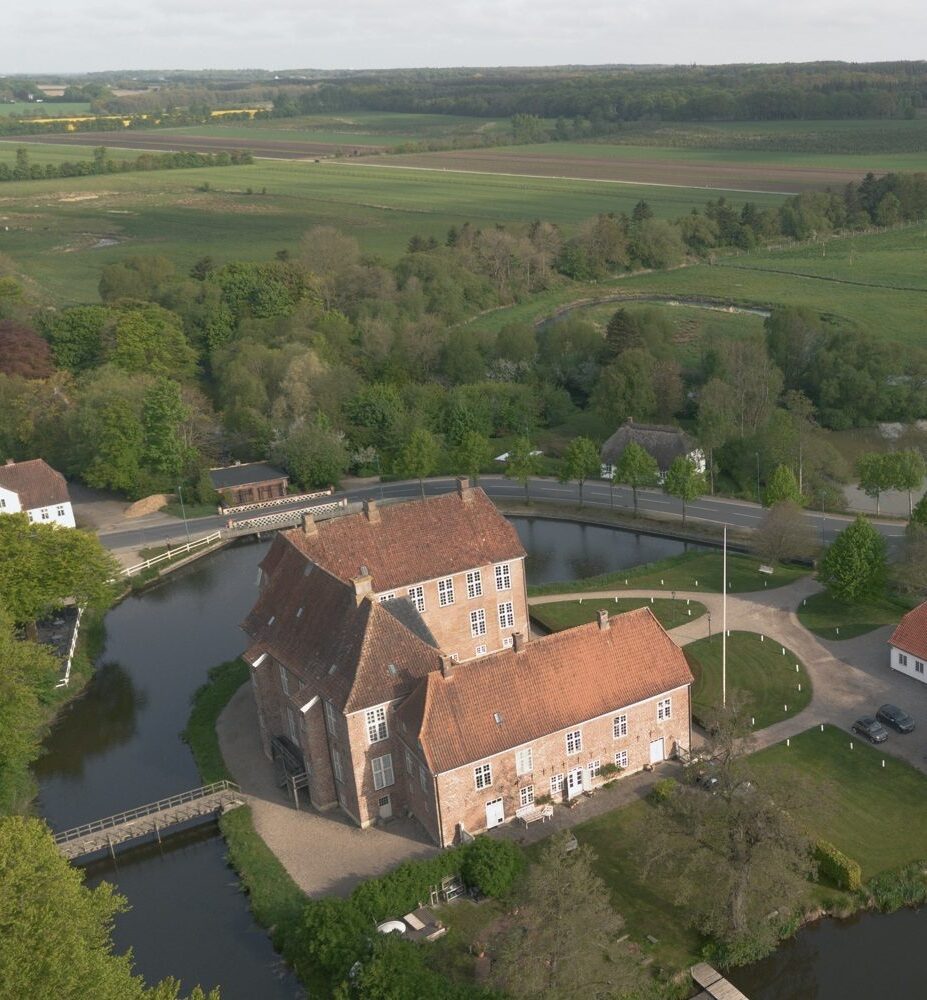 Luftfoto af et historisk murstensslot med røde tage, omgivet af en voldgrav og træer, beliggende i et landligt landskab med marker, veje og yderligere landbrugsbygninger i nærheden.