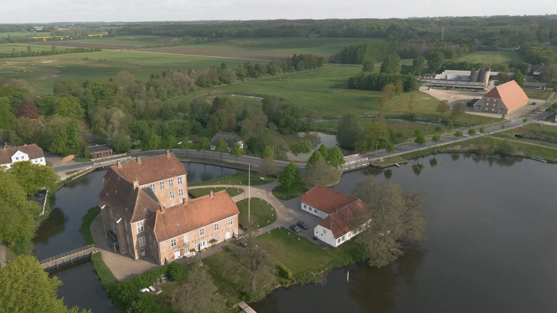Luftfoto af et historisk murstensslot med røde tage, omgivet af en voldgrav og træer, beliggende i et landligt landskab med marker, veje og yderligere landbrugsbygninger i nærheden.