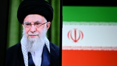 Et portræt af en ældre mand med hvidt skæg, briller og en sort turban vises på en skærm med et sløret iransk flag med det nationale emblem i baggrunden.