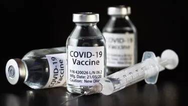 Tre hætteglas mærket "COVID-19 Vaccine" vises, et liggende på siden og to stående, med en sprøjte fyldt med klar væske placeret foran dem, alt sammen på en mørk overflade.
