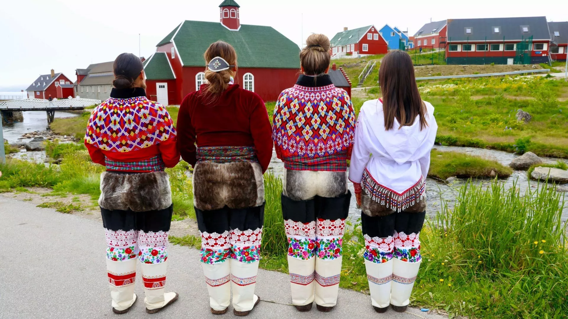Fire kvinder klædt i traditionelt grønlandsk tøj står side om side med ryggen til kameraet og fremviser farverigt perlearbejde, broderet tøj og støvler. Røde, grønne og andre bygninger dukker op i baggrunden.