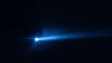 Et lyst objekt i rummet udsender et glødende blåt lys med en lang, smal hale, der strækker sig til højre mod en sort baggrund, og som ligner en komet eller asteroide med et lysende spor.