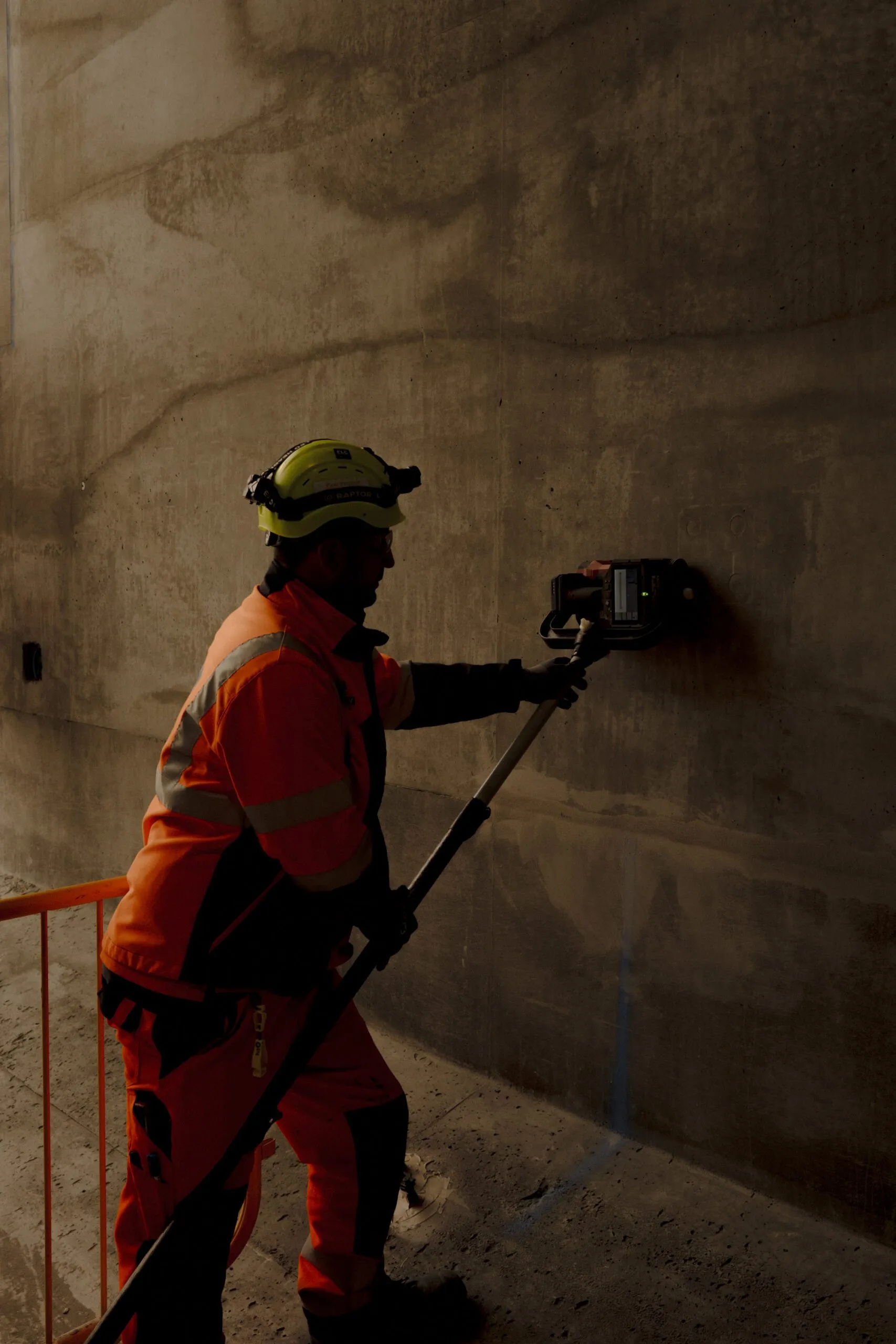 En person i arbejdstøj med høj synlighed og hjelm bruger en scanningsanordning på en betonvæg i et svagt oplyst område, sandsynligvis i forbindelse med bygge- eller inspektionsarbejde.