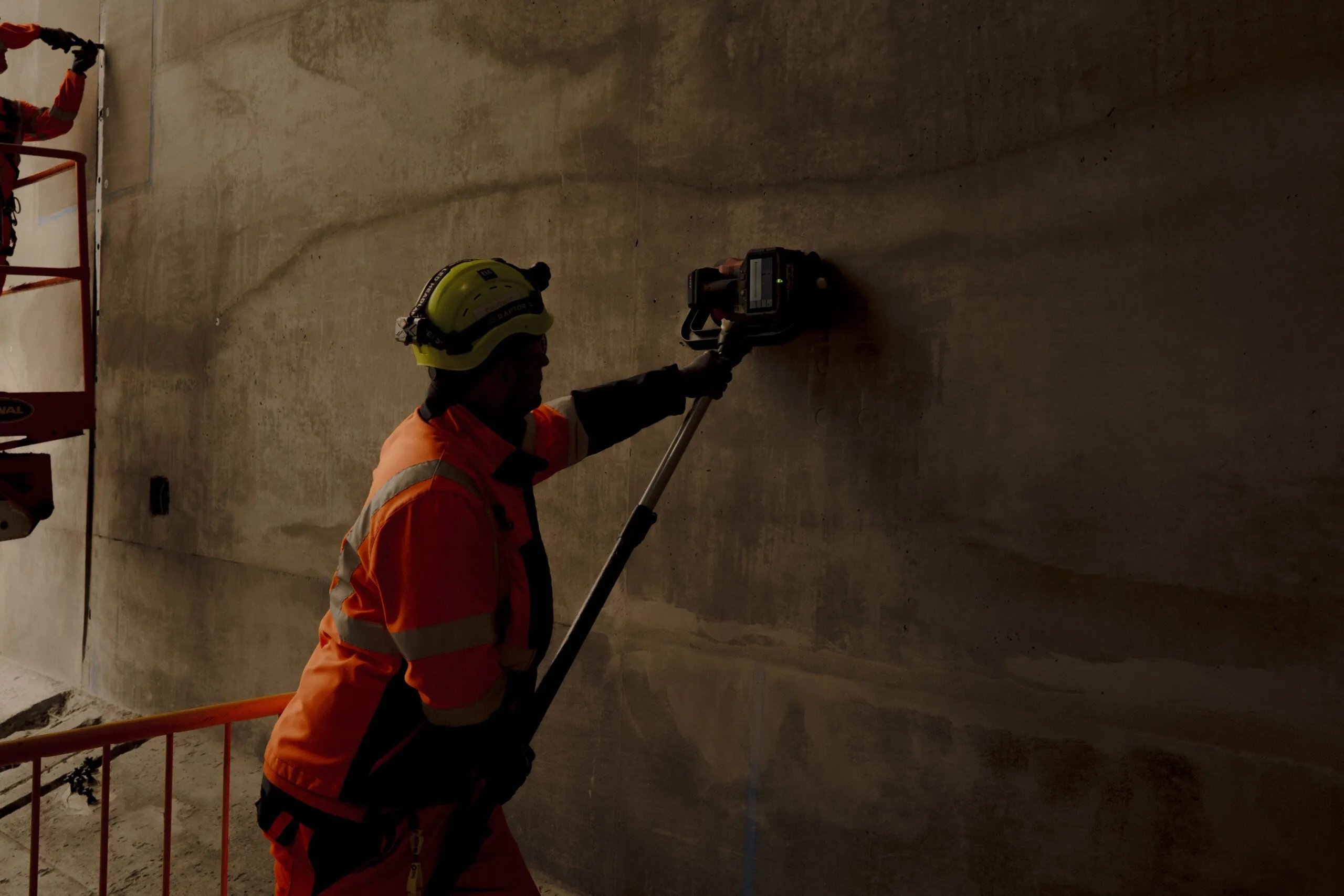 En bygningsarbejder i jakke og hjelm med høj synlighed bruger et værktøj på en betonvæg i et svagt oplyst område; en anden arbejder er hævet op på en platform i baggrunden.